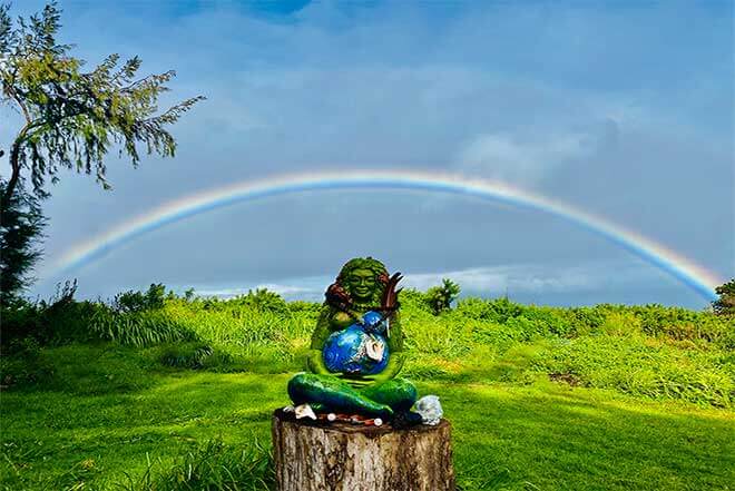 Female deity sitting on a tree stump outdoors with a full rainbow in the background at Earthsong Foundation Hawaii